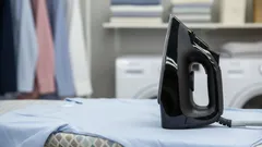 Černá žehlička na žehlícím prkně v prádelně, v pozadí pračka a košile na ramínku.