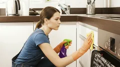 Žena v rukavicích uklízí kuchyňskou linku pomocí čisticího prostředku a hadříku.