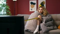Žena sleduje televizi na gauči, zabalená v dece, domácí pohoda v obývacím pokoji.