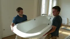 Dva muži stěhují vanu v prázdném bytě během rekonstrukce nebo zařizování.