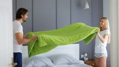Mladý pár společně povléká postel zeleným povlečením v moderní ložnici.