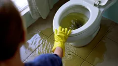 Zablokovaná toaleta, přetékající voda na podlaze, osoba v gumových rukavicích čistí záchod.